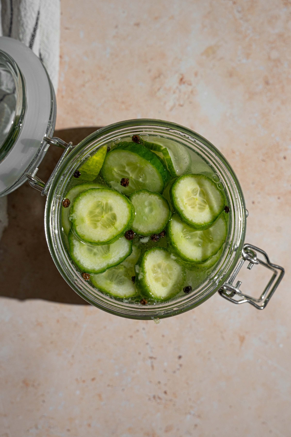 An open jar with sliced cucumbers pickling in a vinegar mixture with black peppercorns. The jar is on a tan counter.