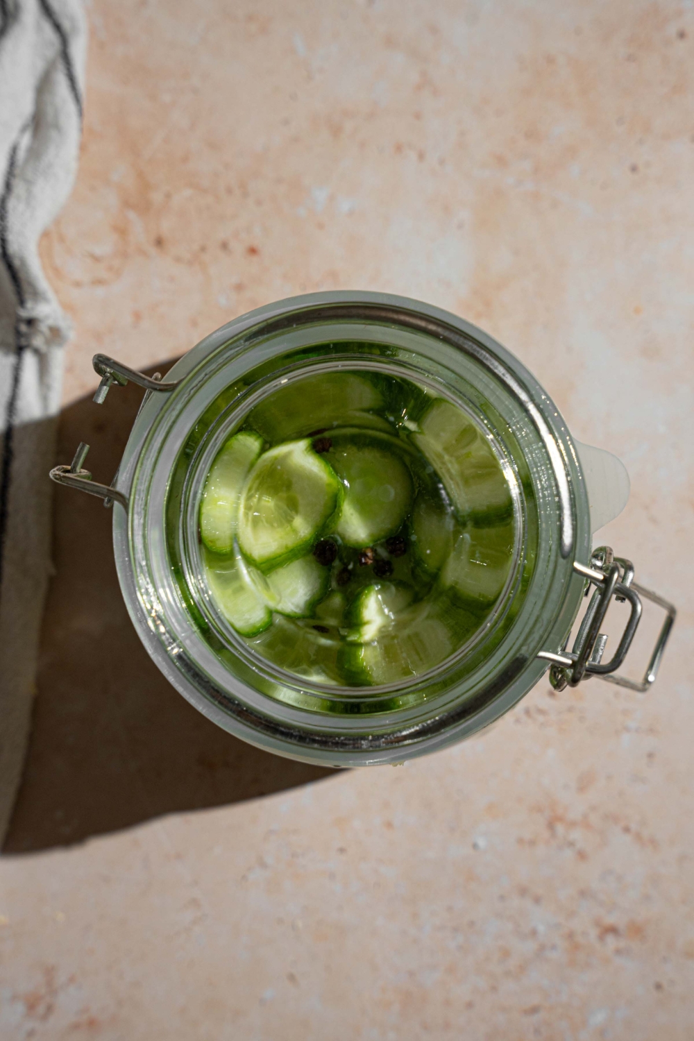 A sealed jar with sliced cucumbers pickling in a vinegar mixture with black peppercorns. The jar is on a tan counter.