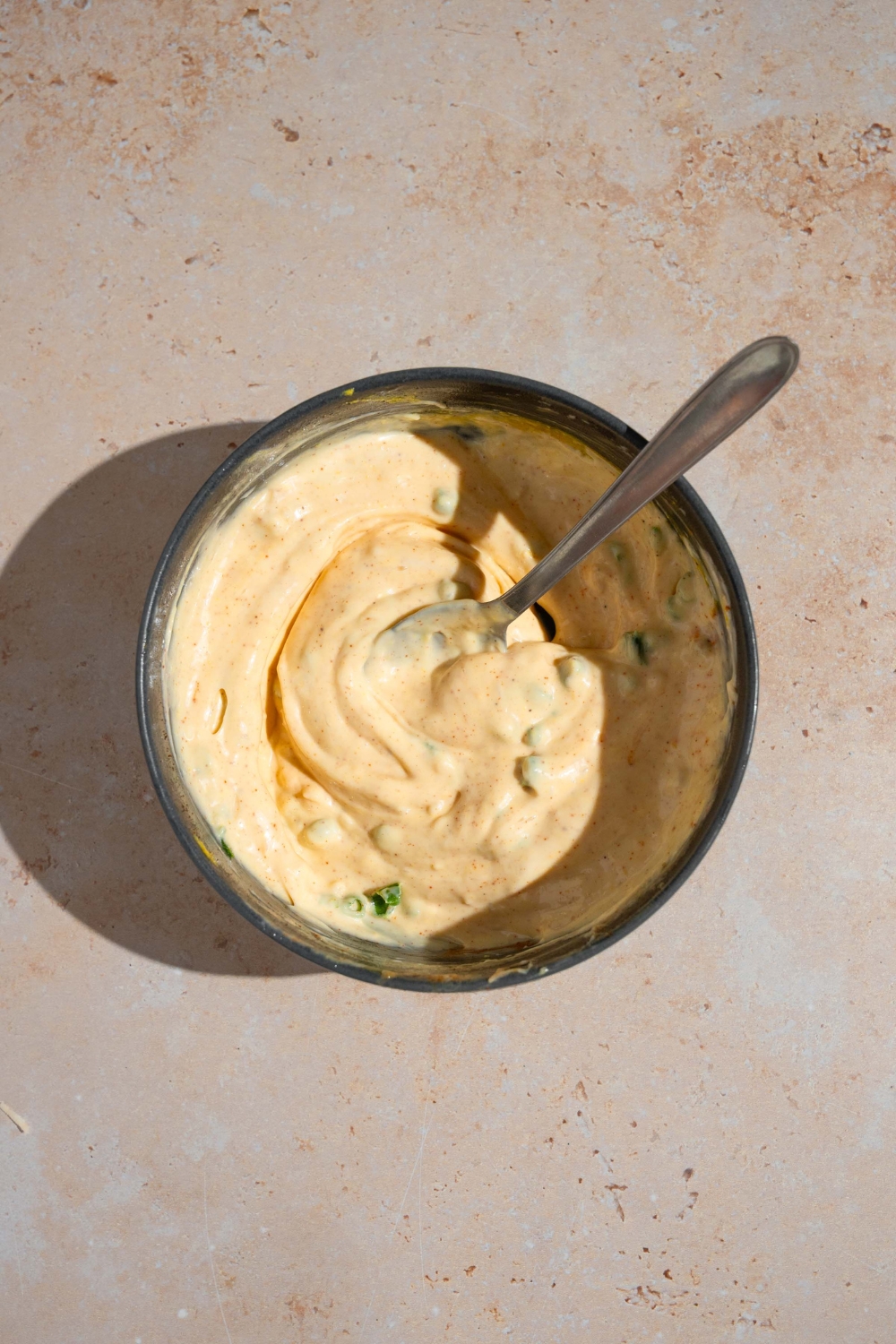 A small bowl with a spoon stirring ingredients to make remoulade sauce. The bowl is on a tan counter.