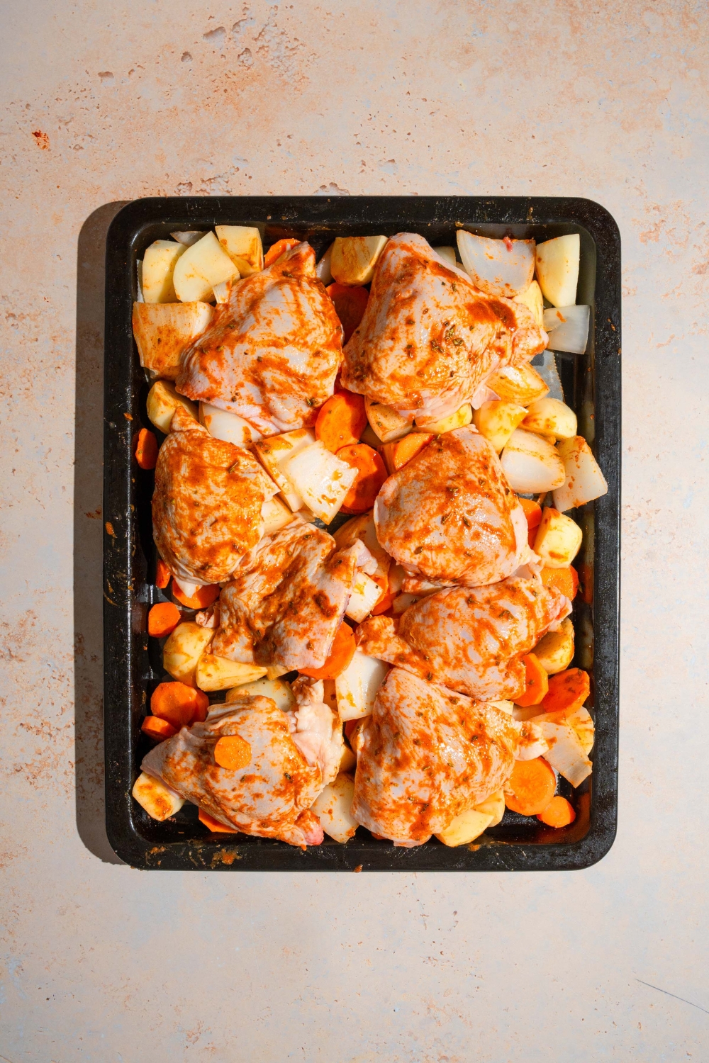A baking sheet with marinated chicken thighs with sliced carrots, potatoes, and onions. The sheet is on a tan counter.