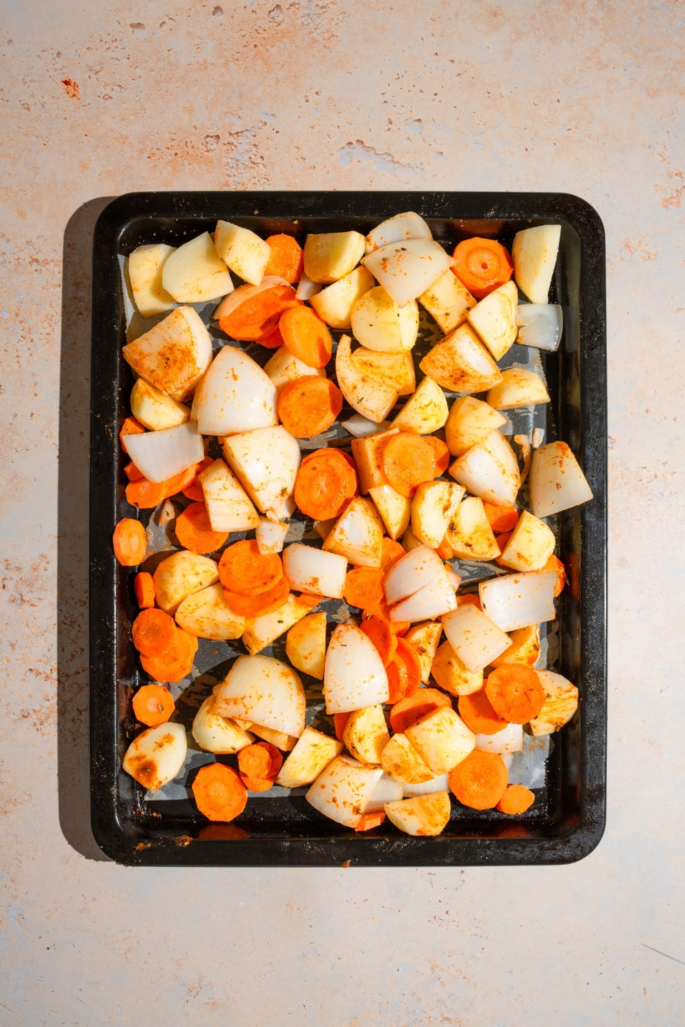 A baking sheet lined with parchment paper with seasoned sliced carrots, onions, and potatoes. The sheet is on a tan counter.