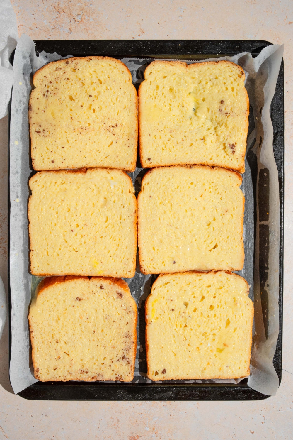A baking sheet lined with parchment paper with several slices of uncooked french toast. The sheet is on a tan counter.