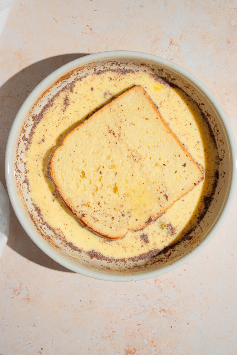A bowl with a slice of brioche bread soaking in an egg and milk mixture. The bowl is on a tan counter.