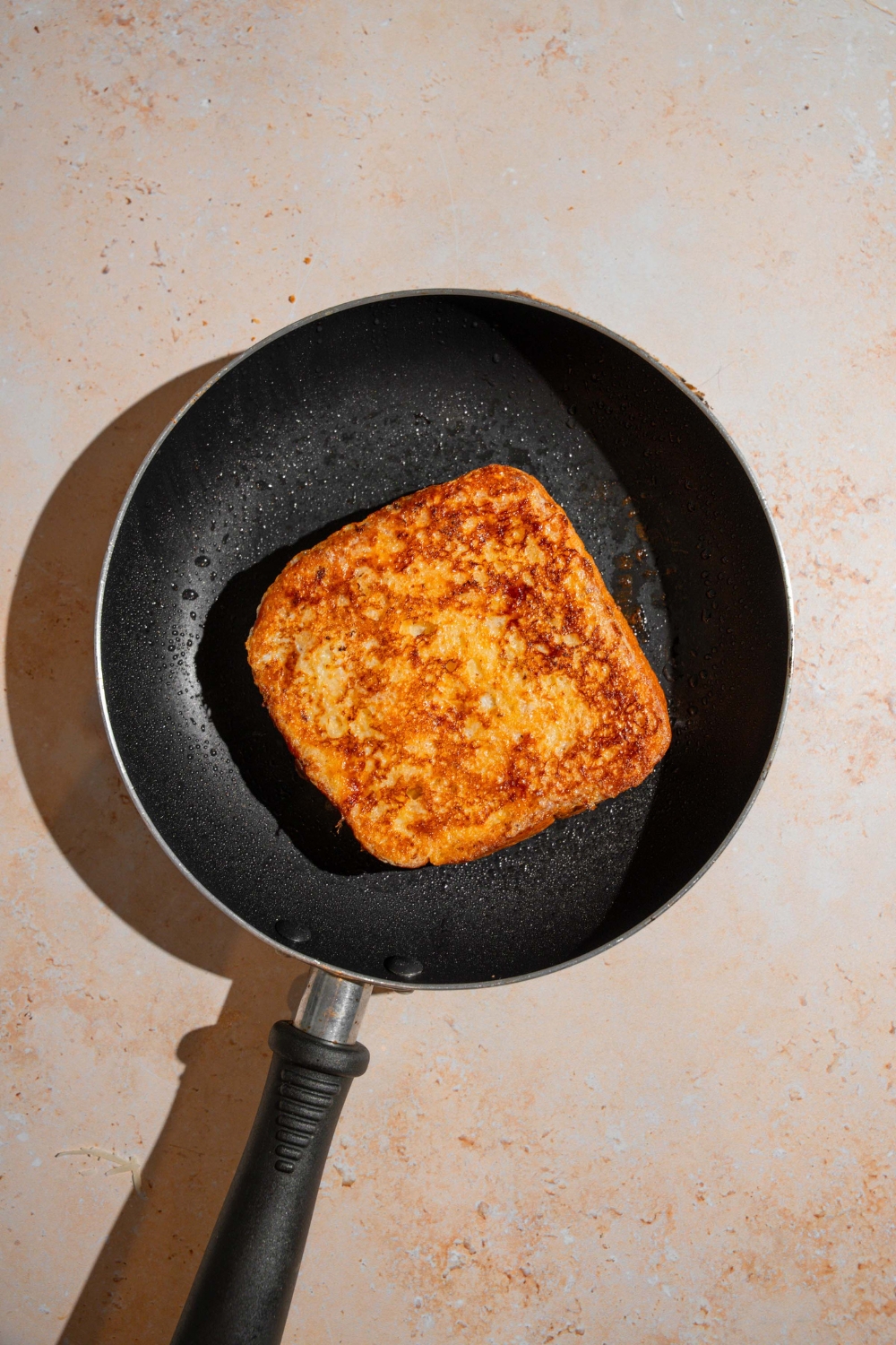 A skillet with a slice of cooked French toast. The skillet is on a tan counter.