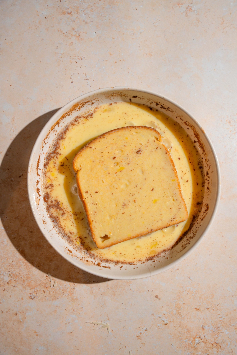 A white bowl with a piece of brioche bread soaking in a french toast milk mixture. The bowl is on a tan counter.