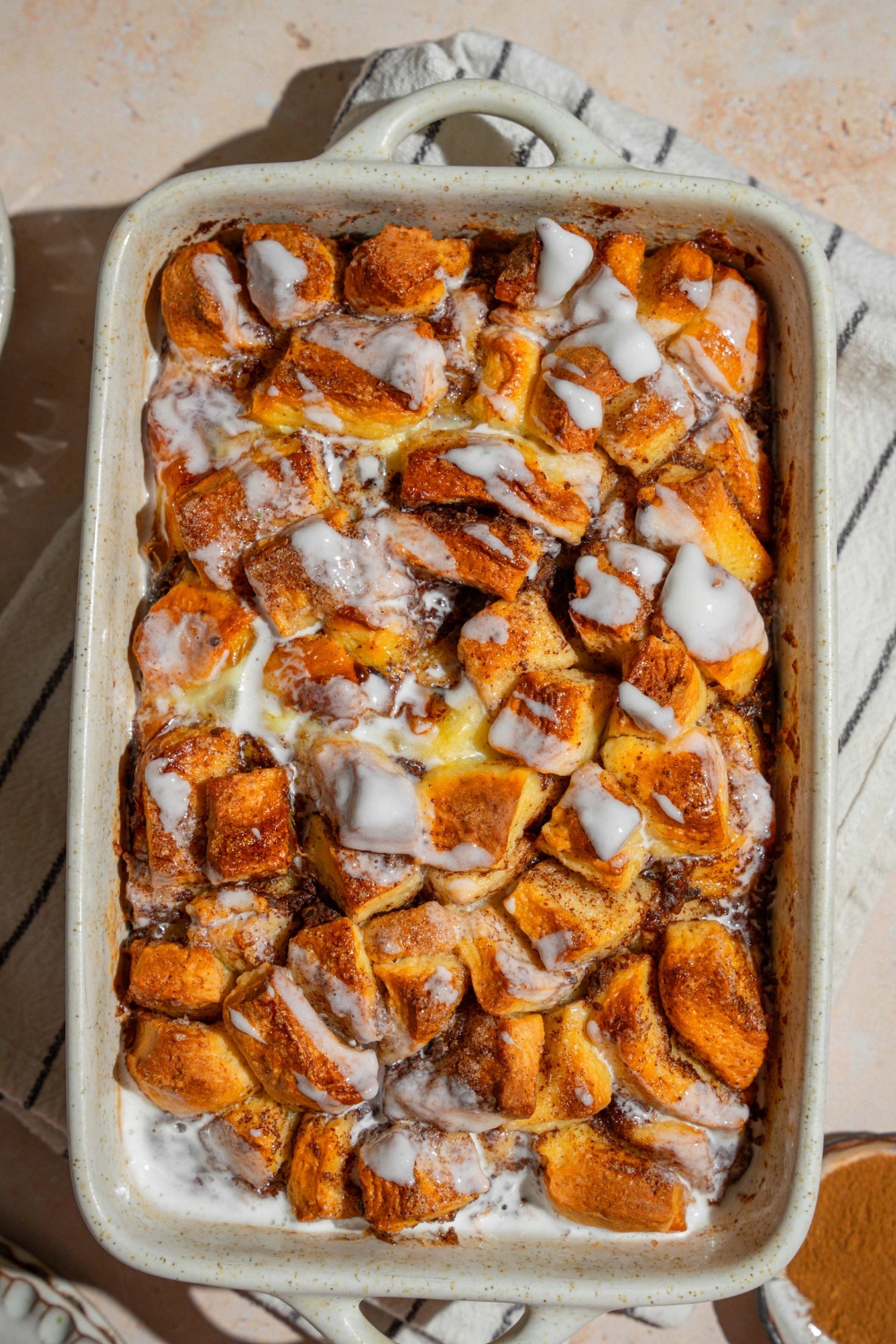 A baking dish with cinnamon roll french toast drizzled with icing. The dish is on a tan counter on a white striped napkin.