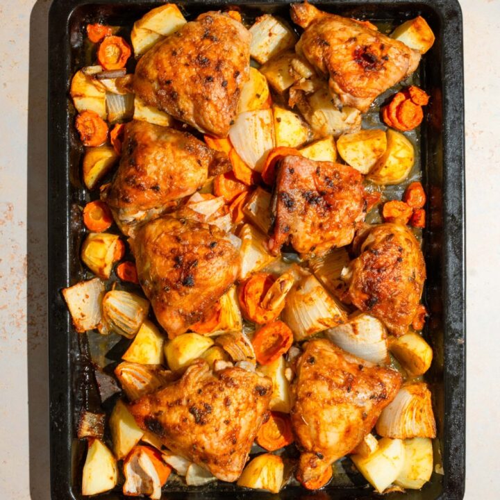 A baking sheet lined with parchment paper with roasted chicken thighs, carrots, potatoes, and onions. The sheet is on a tan counter.