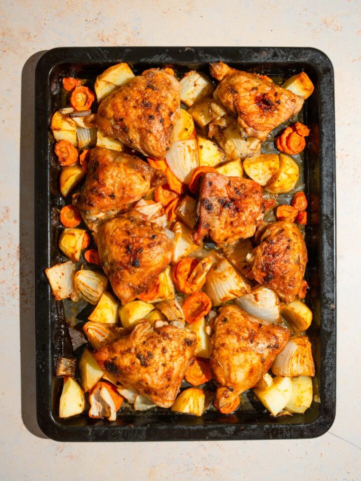 A baking sheet lined with parchment paper with roasted chicken thighs, carrots, potatoes, and onions. The sheet is on a tan counter.