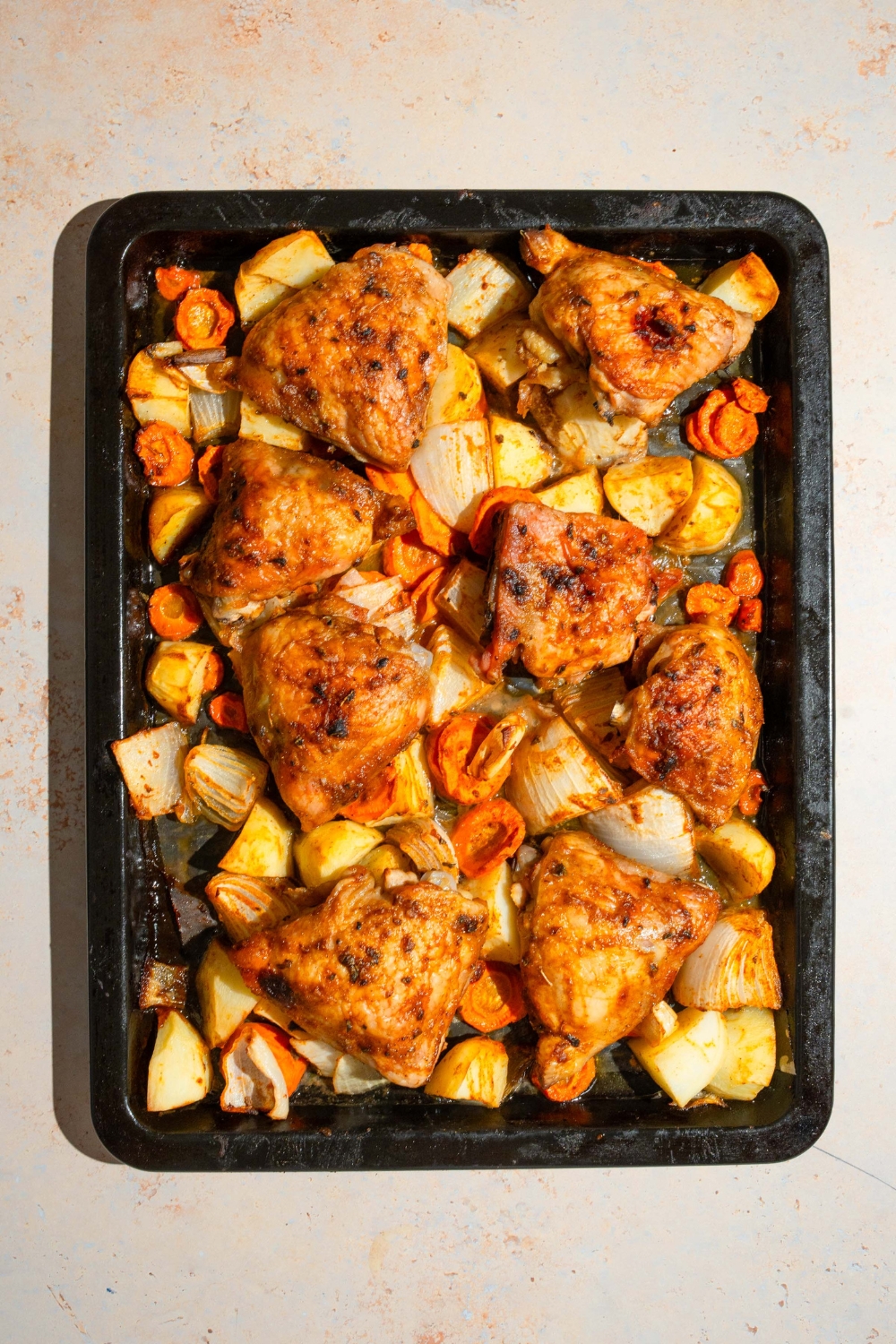 A baking sheet lined with parchment paper with roasted chicken thighs, carrots, potatoes, and onions. The sheet is on a tan counter.