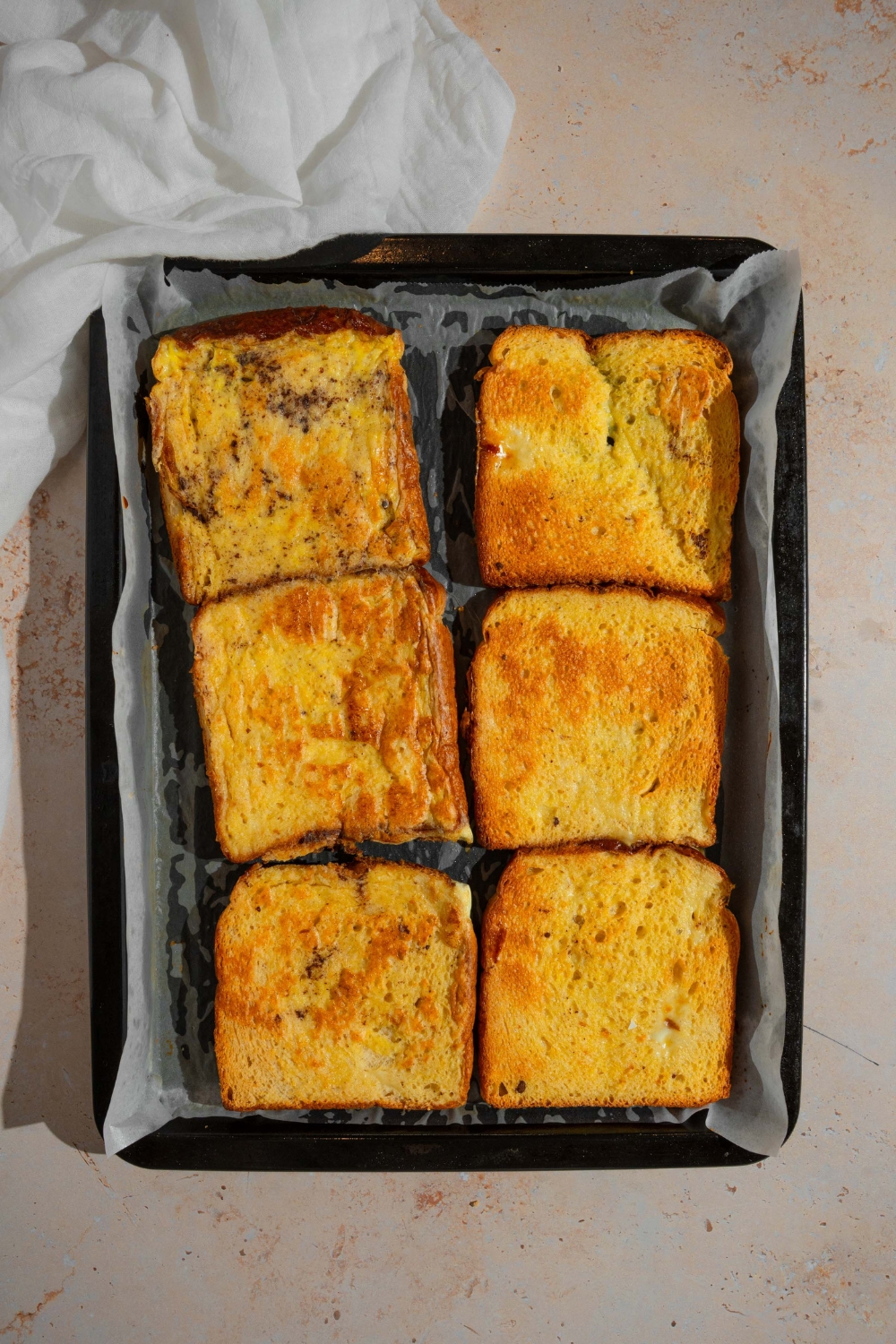 A baking sheet lined with parchment paper with several pieces of baked french toast. The sheet is on a tan counter.