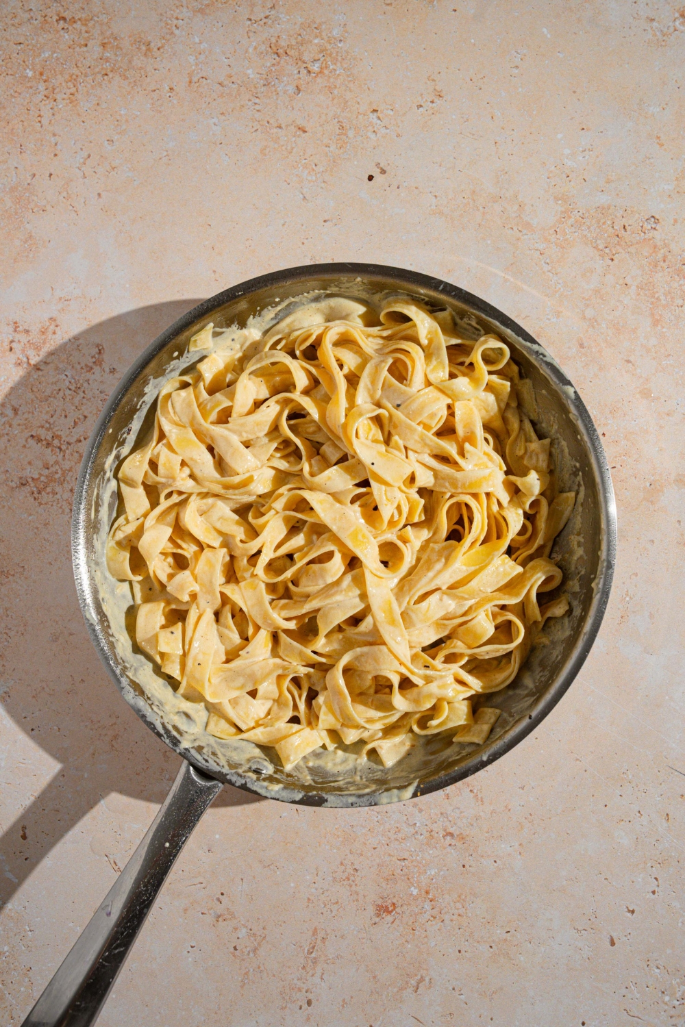 A skillet with fettuccine pasta tossed in alfredo sauce. The skillet is on a tan counter.