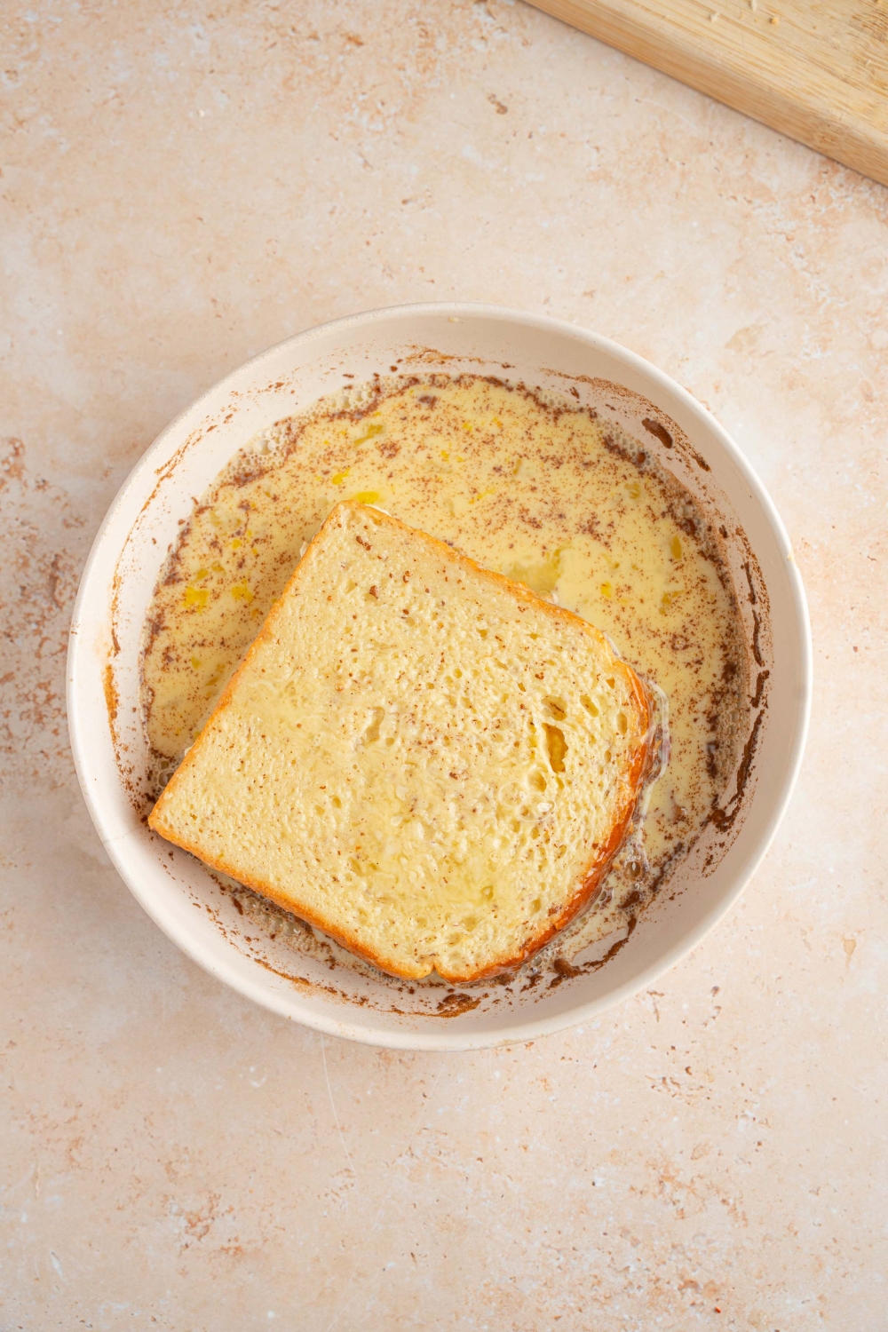 A white bowl with a milk mixture with a slice of bricohe bread soaking in the mixture. The bowl is on a tan counter.