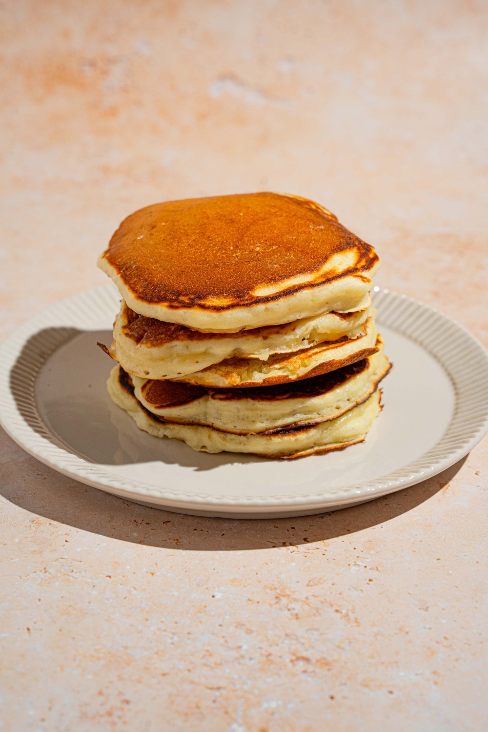 A white plate with a stack of sweet cream pancakes. The plate is on a tan counter.