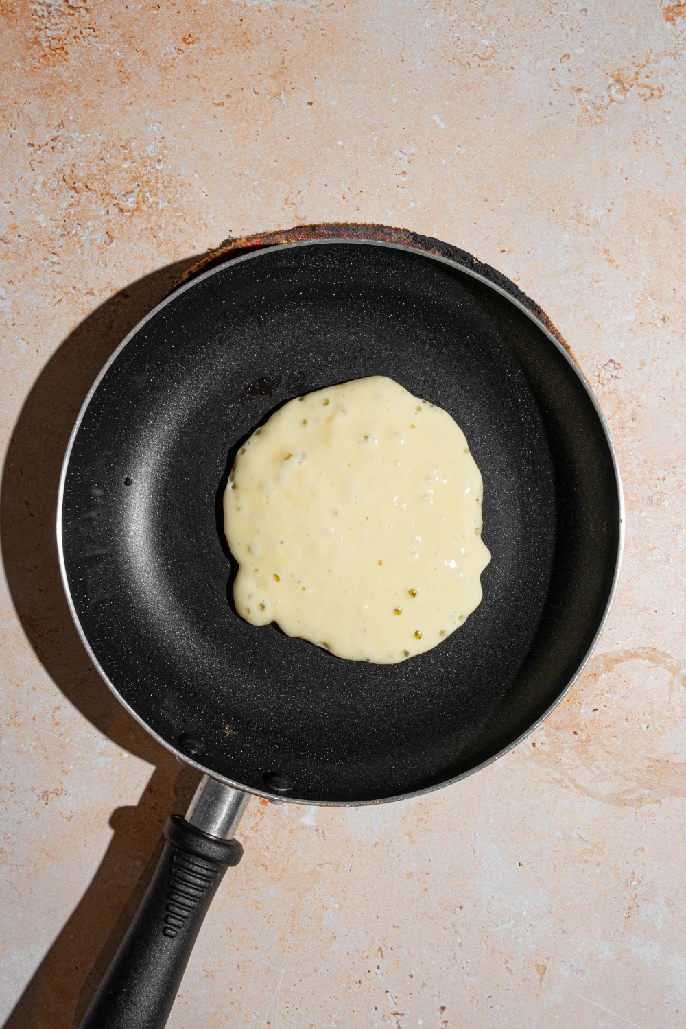 A skillet with an uncooked sweet cream pancakes. The skillet is on a tan counter.
