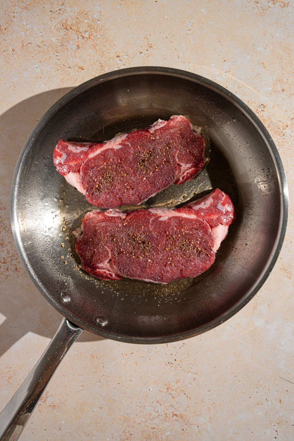 A skillet with two seasoned uncooked steaks in butter. The skillet is on a tan counter.
