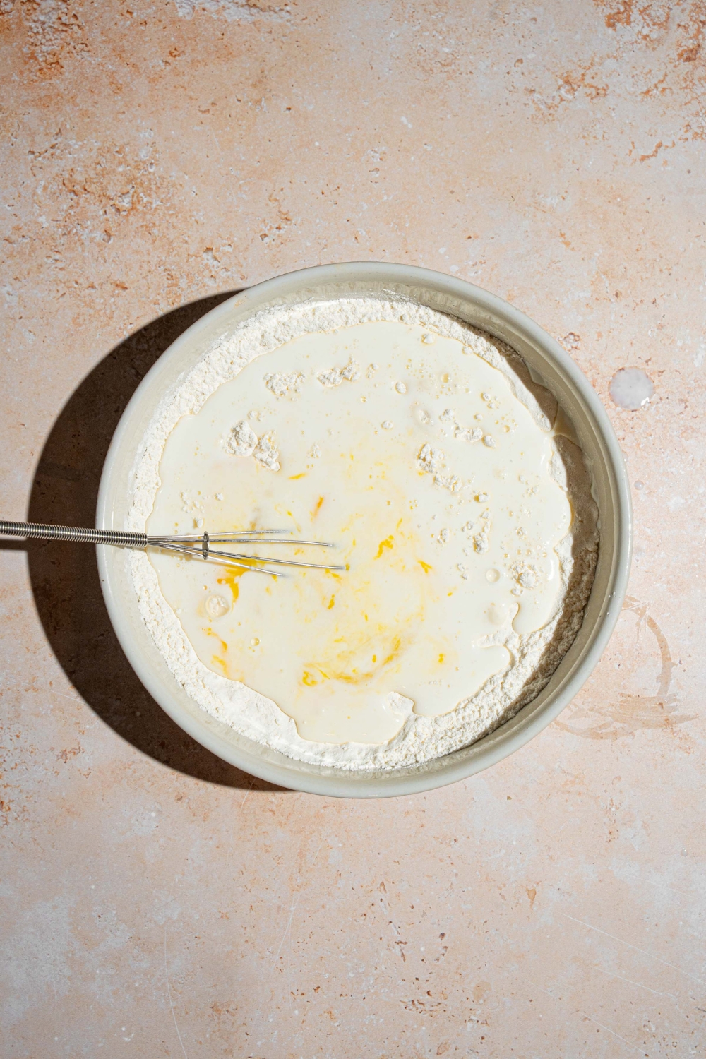 A white bowl with a whisk mixing wet and dry ingredients for sweet cream pancakes ingredients. The bowl is on a tan counter.