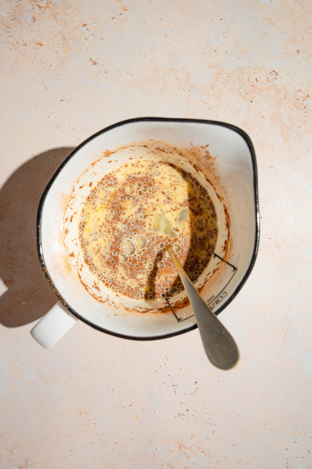 A ceramic pitcher with a spoon mixing an egg mixture with milk, cinnamon, and vanilla. The pitcher is on a tan counter.