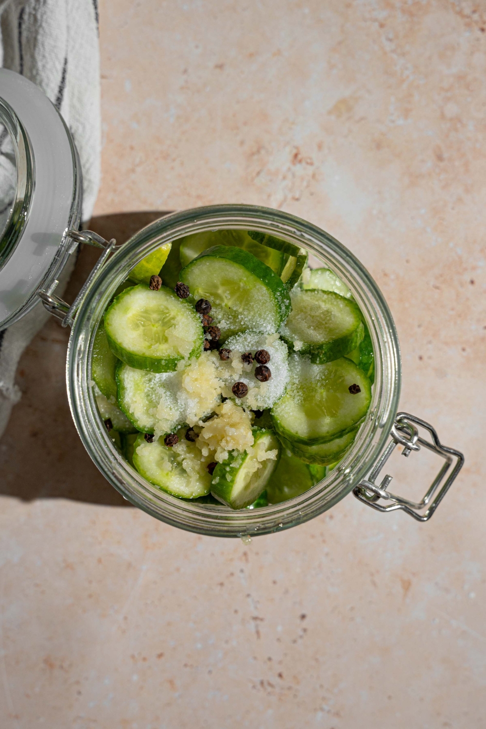 An open jar with sliced cucumbers in a vinegar mixture topped with salt, black peppercorns, and garlic. The jar is on a tan counter.