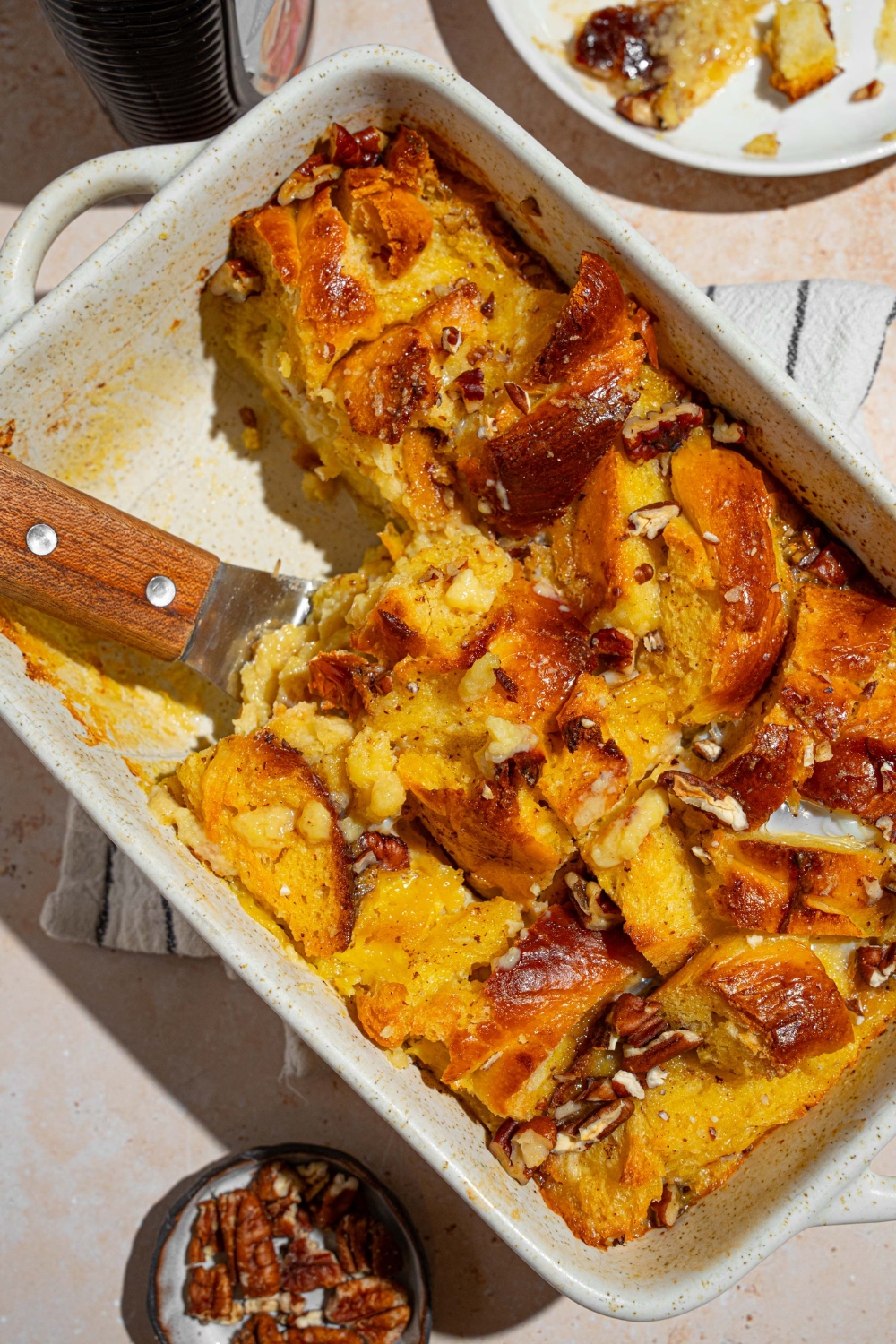 A baking dish with challah french toast with a spatula removing a piece. The dish is on a tan counter with a white striped napkin and small plate of pecans.
