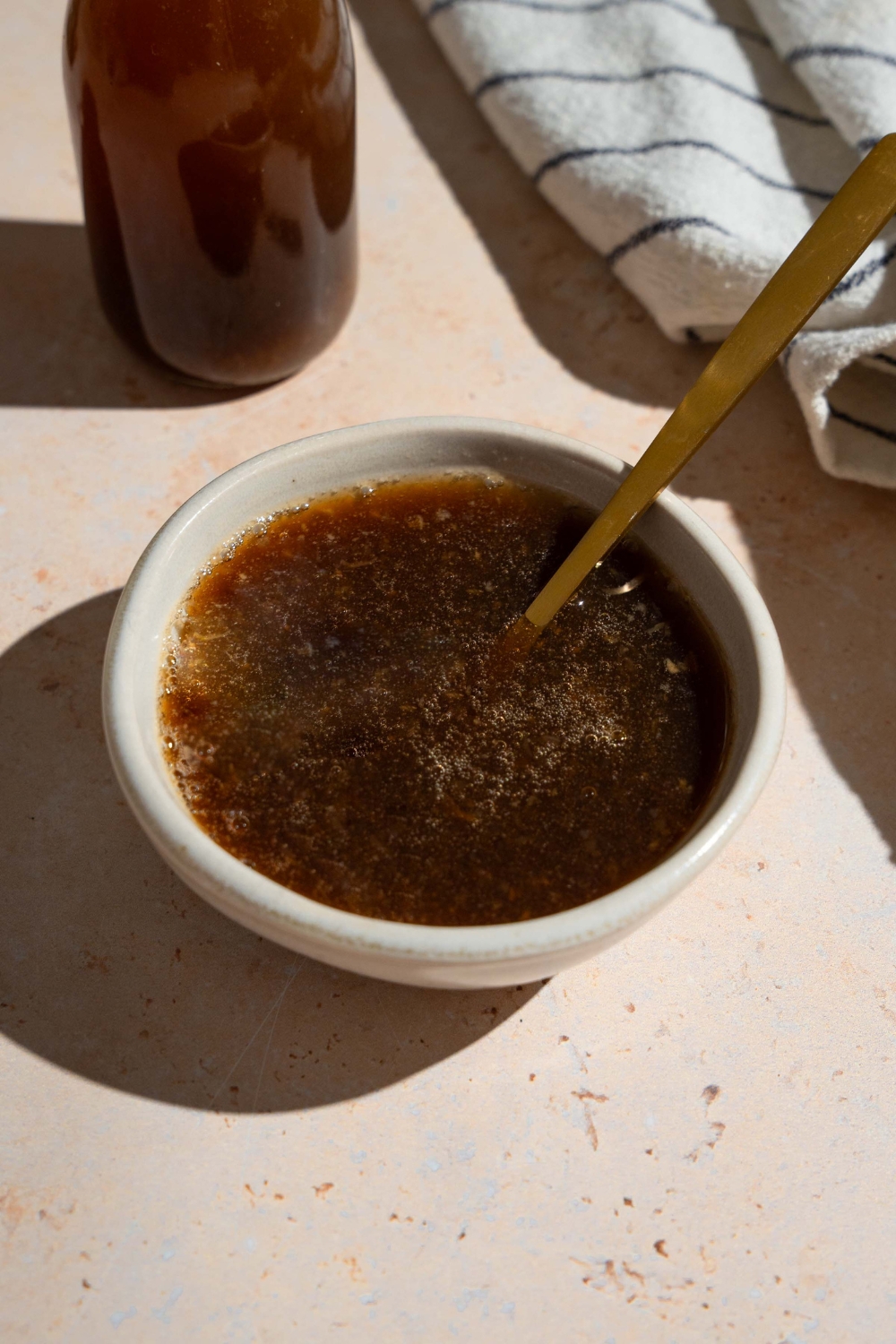 A bowl of oyster sauce with a spoon. The bowl is on a tan counter with a white striped napkin and jar of sauce.