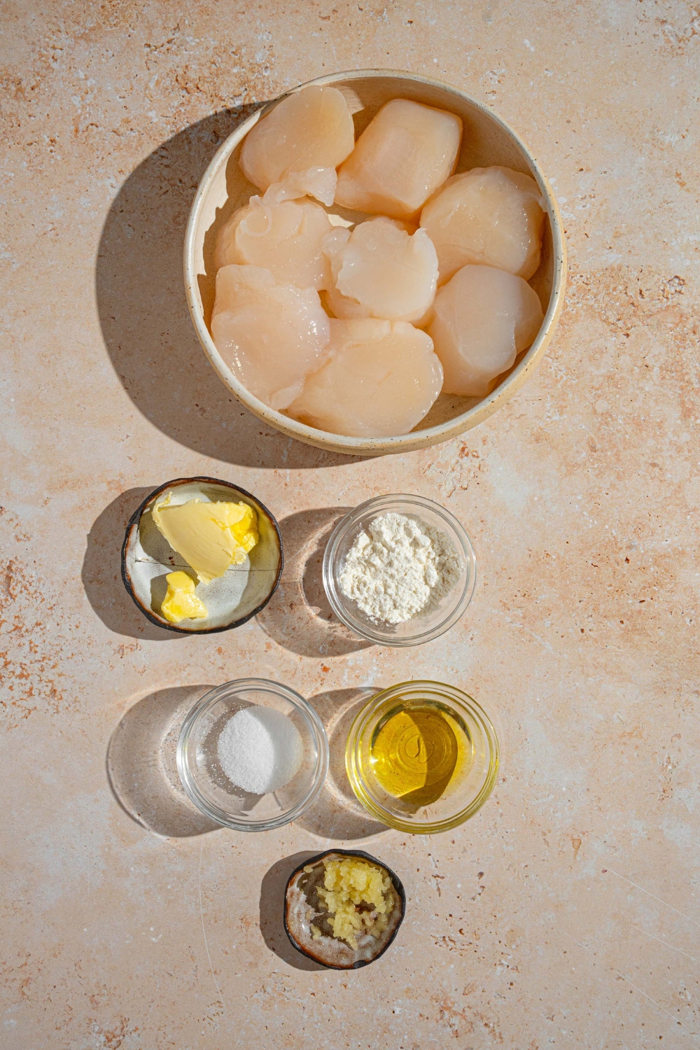 An overhead shot of several bowls in various sizes containing ingredients to make pan seared scallops including frozen scallops, garlic, flour, butter, oil, and seasonings.