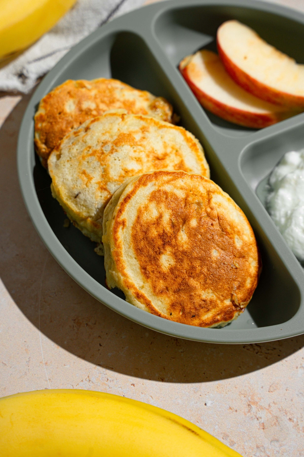 A kids plate with three 3 ingredient banana pancakes with a side of sliced apples and cottage cheese. The plate is on a tan counter with a white striped napkin and bananas.