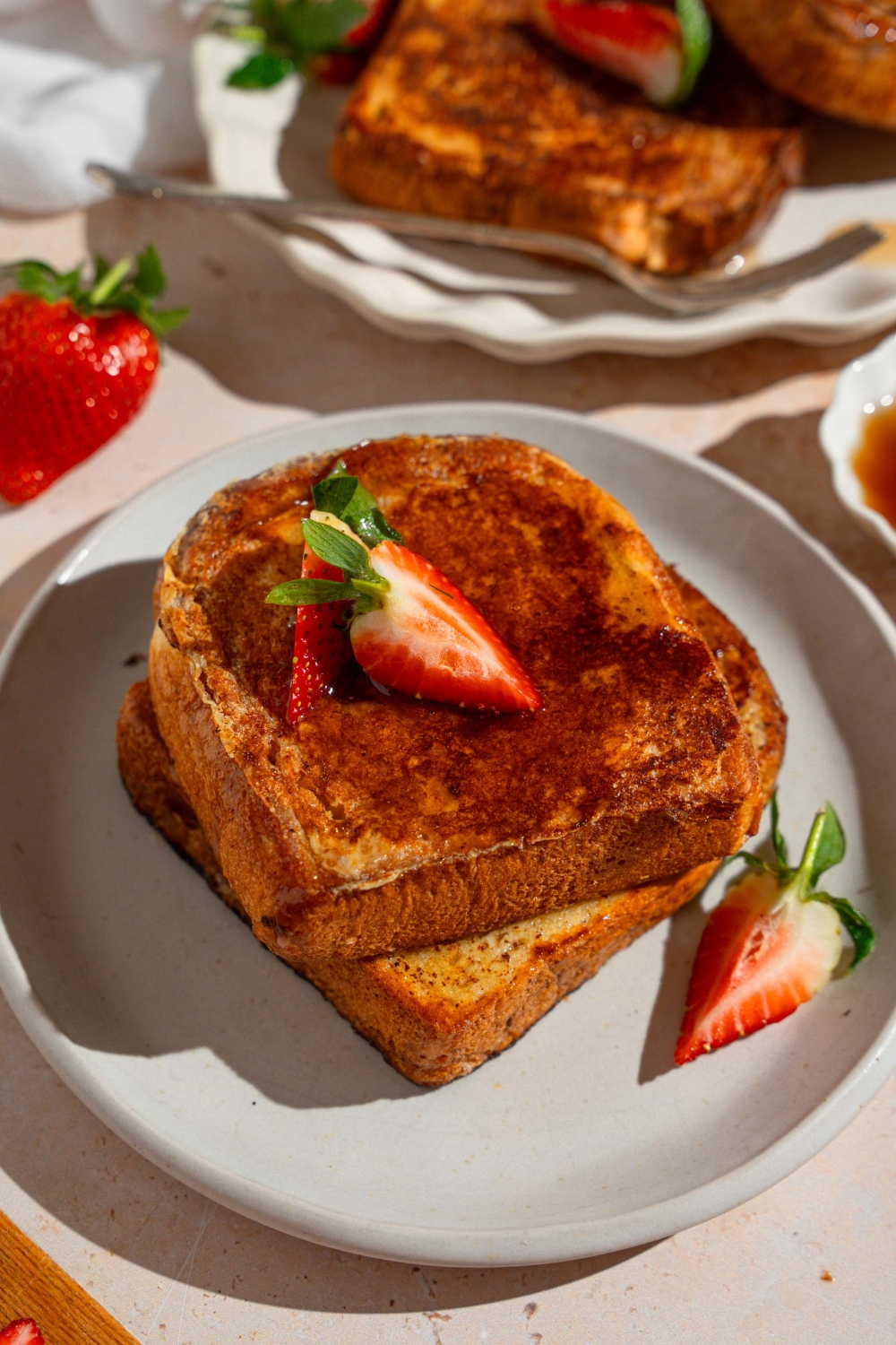 A white plate with slices of French toast drizzled with maple syrup and garnished with sliced strawberries. The plate is on a tan counter with a platter of french toast.