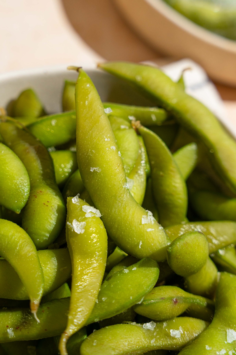A bowl of cooked edamame seasoned with salt. The bowl is on a tan counter with a bowl of frozen edamame.
