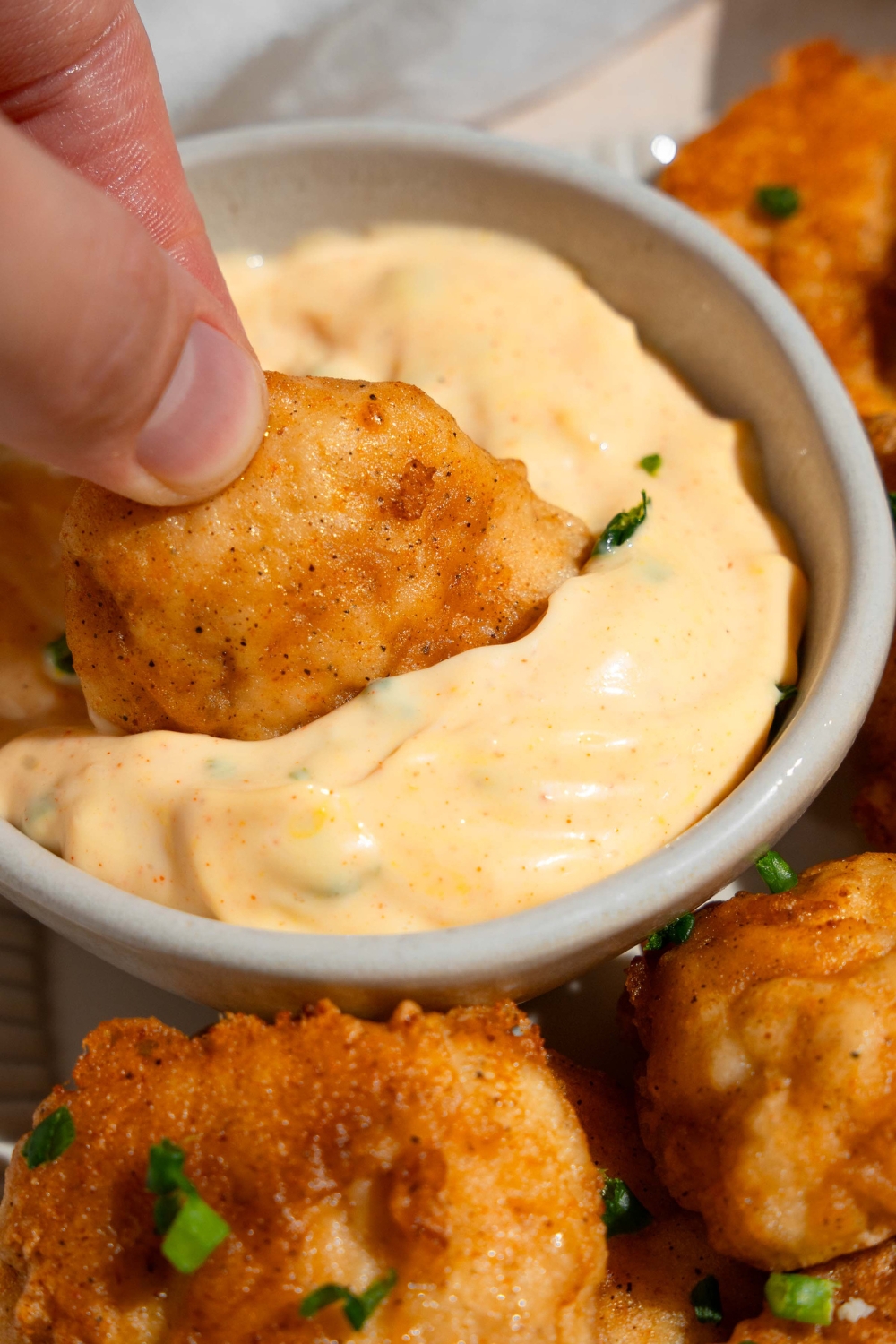 A hand dipping a chicken nugget into a bowl with remoulade sauce. The bowl is on a plate of chicken nuggets.