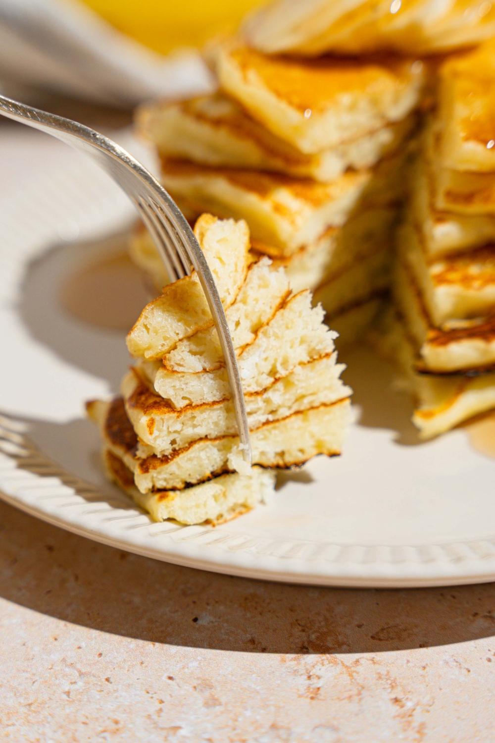 A close up of a fork with a bite taken from a stack of pancakes. The fork is on a white plate with pancakes.