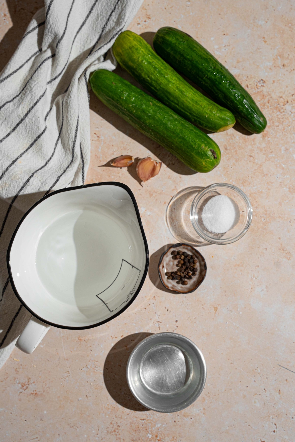 An overhead shot of several bowls in various sizes containing ingredients to make quick pickles including cucumbers, vinegar, salt, garlic, black peppercorns, and water.