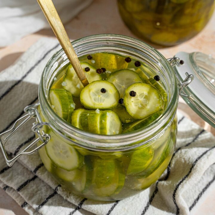 A jar with quick pickles in juices. There is a fork in the jar. The jar is on a white striped napkin on a tan counter with a small plate of sliced pickles.
