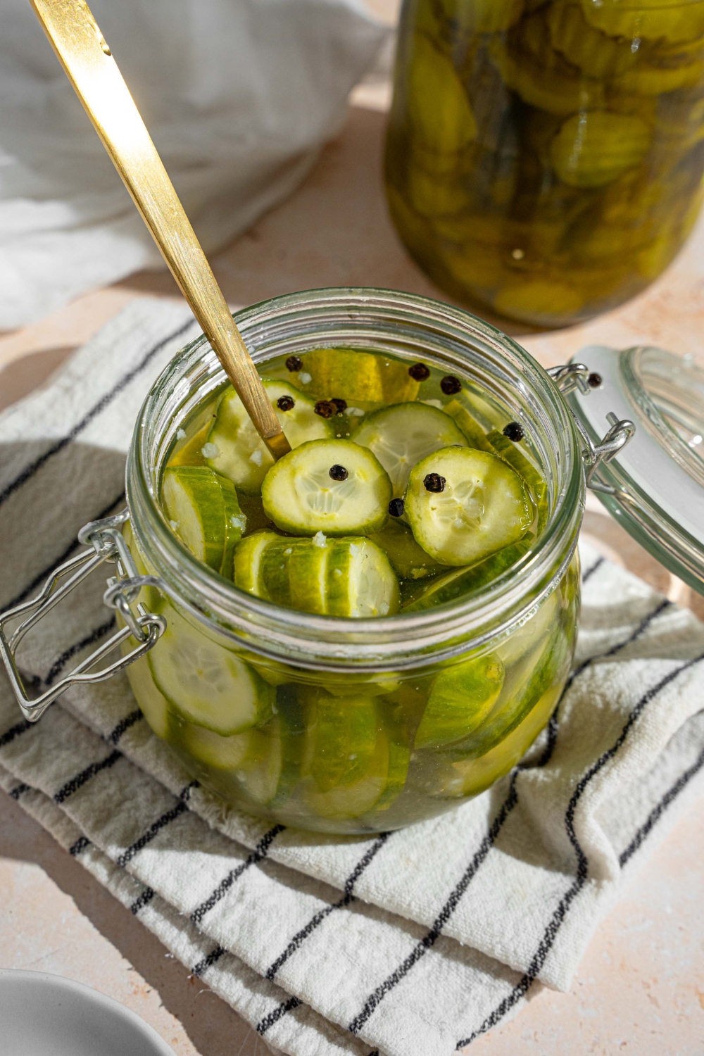 A jar with quick pickles in juices. There is a fork in the jar. The jar is on a white striped napkin on a tan counter with a small plate of sliced pickles.