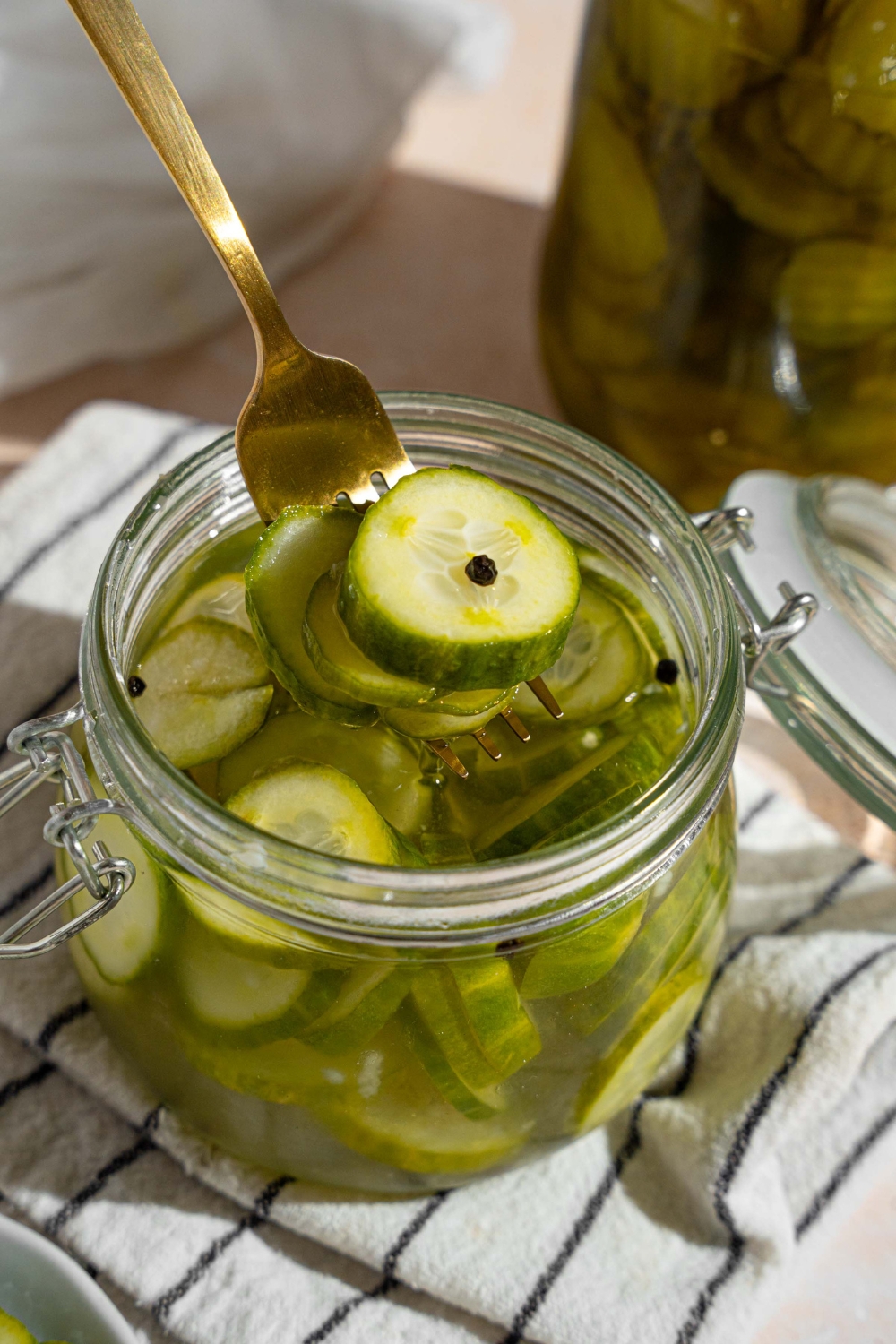 A jar with quick pickles in juices. There is a fork taking pickles from the jar. The jar is on a white striped napkin on a tan counter with a small plate of sliced pickles.
