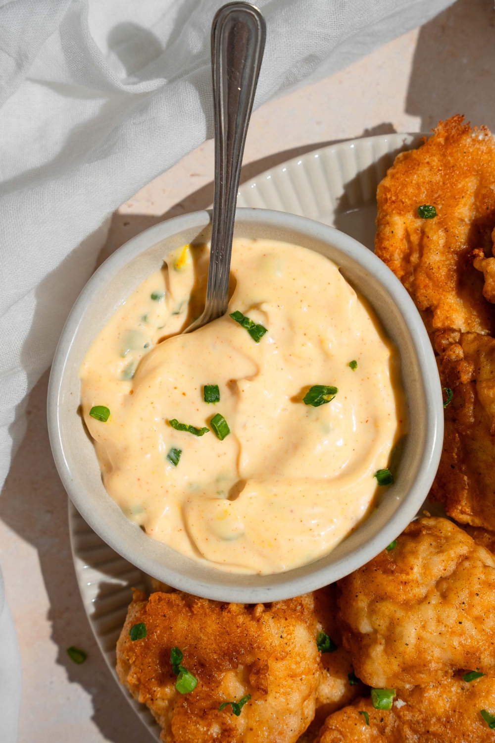 A ramekin of remoulade sauce garnished with chopped chives and served with a spoon. The bowl is on a plate of chicken nuggets on a tan counter with a white cloth napkin.