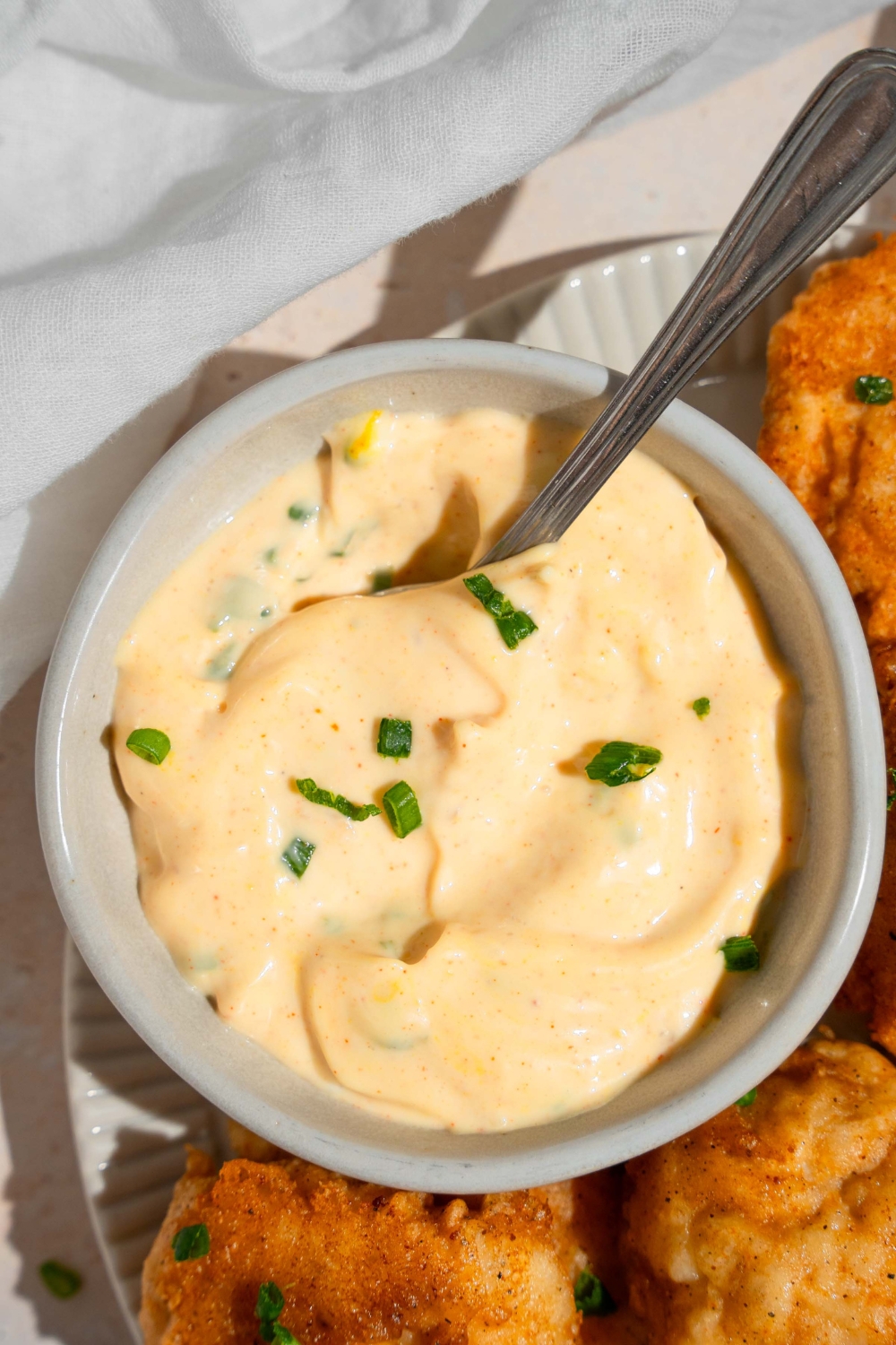 A ramekin of remoulade sauce garnished with chopped chives and served with a spoon. The bowl is on a plate of chicken nuggets on a tan counter with a white cloth napkin.
