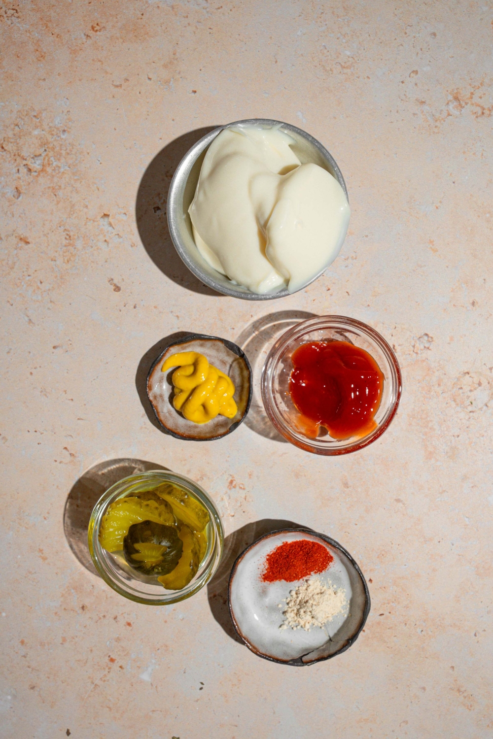 An overhead shot of several bowls in various sizes containing ingredients to make Shake Shack sauce including mayo, ketchup, mustard, pickles, and seasonings.