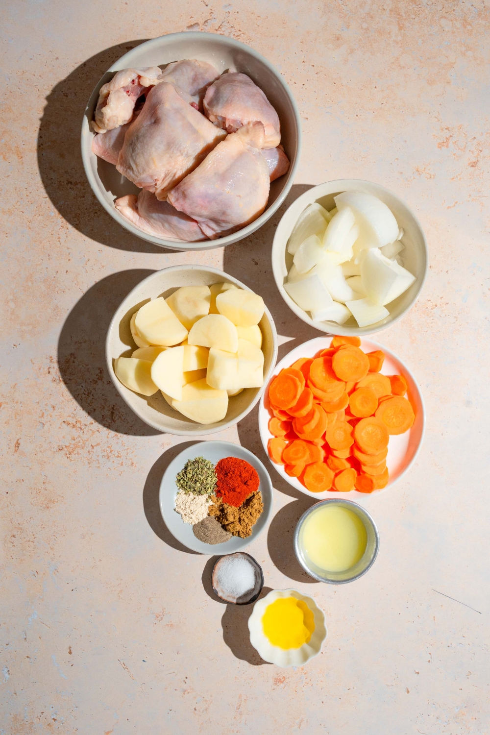 An overhead shot of several bowls in various sizes containing ingredients to make sheet pan chicken thighs including chicken thighs, carrots, potatoes, onions, lime juice, oil, and seasonings.