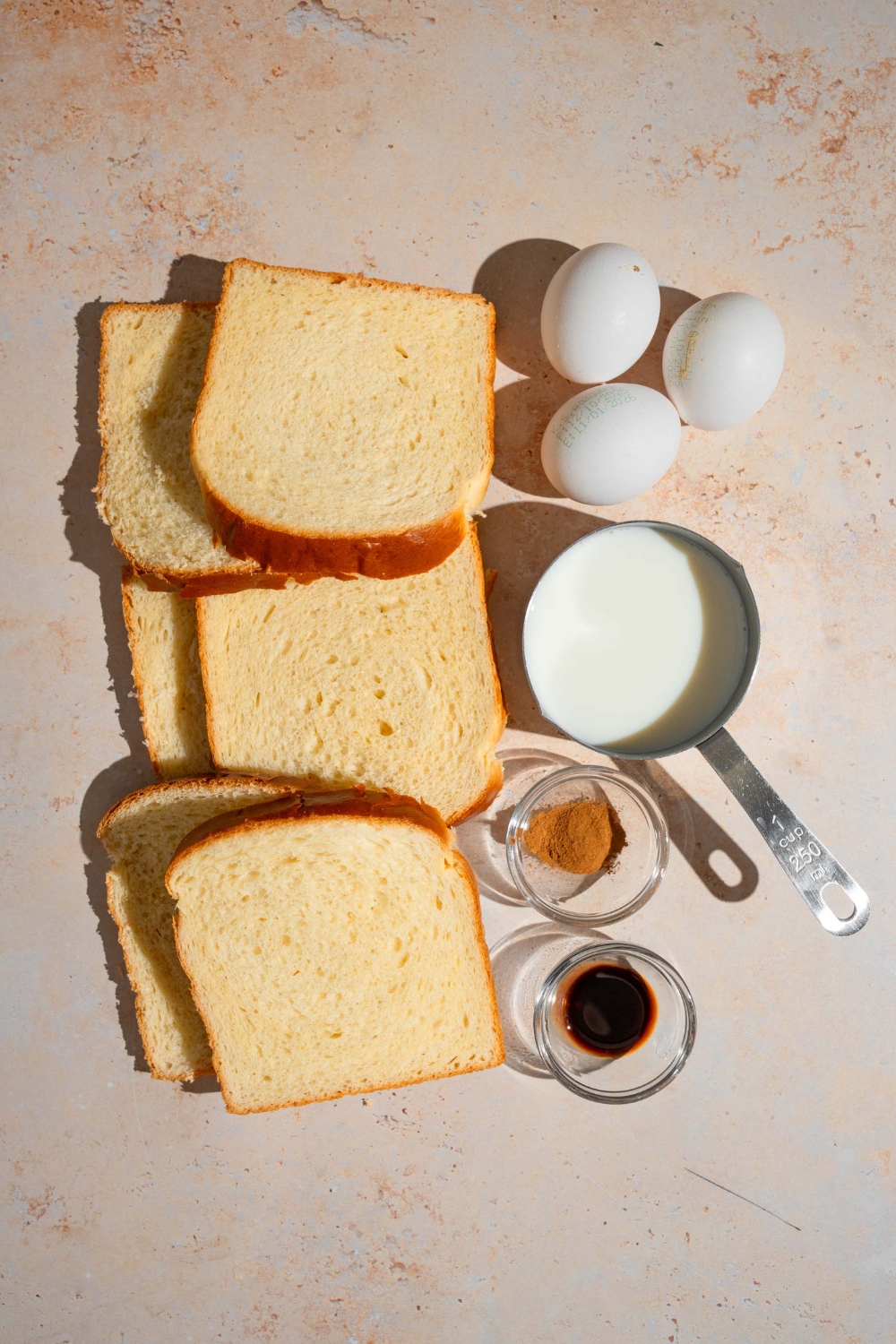 An overhead shot of several bowls in various sizes containing ingredients to make sheet pan french toast including slices of brioche bread, eggs, milk, vanilla, and cinnamon.