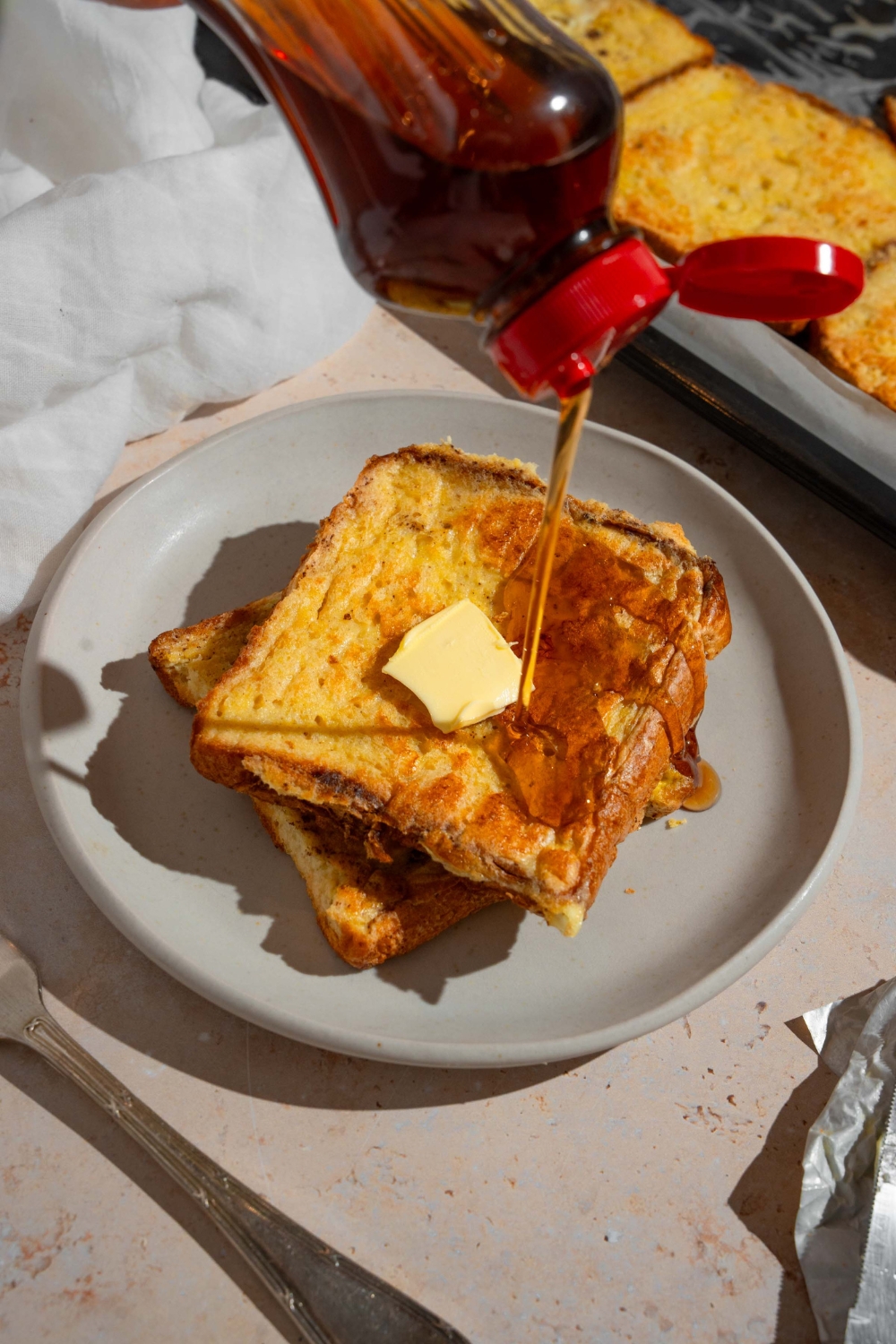 A plate with two slices of sheet pan french toast topped with butter. Syrup is being poured on the plate. The plate is on a tan counter with a fork and sheet pan of french toast.