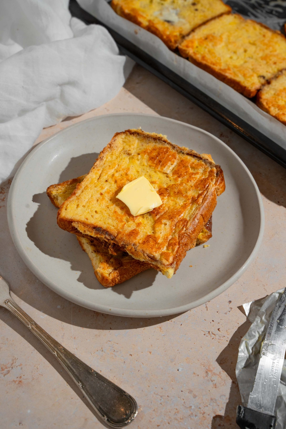 A plate with two slices of sheet pan french toast topped with butter. The plate is on a tan counter with a fork and sheet pan of french toast.