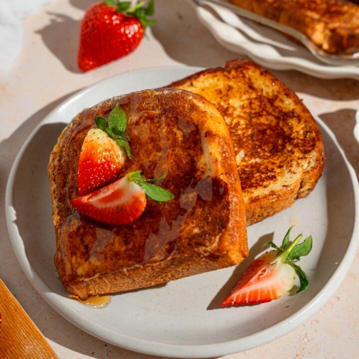 A white plate with slices of French toast drizzled with maple syrup and garnished with sliced strawberries. The plate is on a tan counter with a platter of french toast.