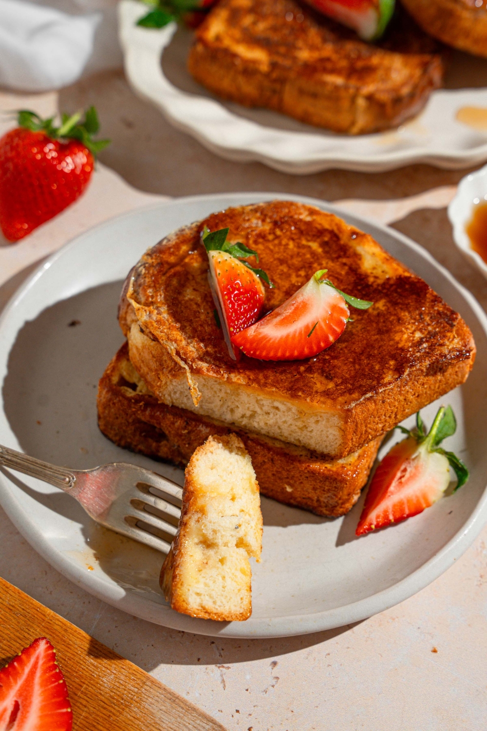 A white plate with slices of French toast drizzled with maple syrup and garnished with sliced strawberries. A fork has taken a bite of toast. The plate is on a tan counter with a platter of french toast.