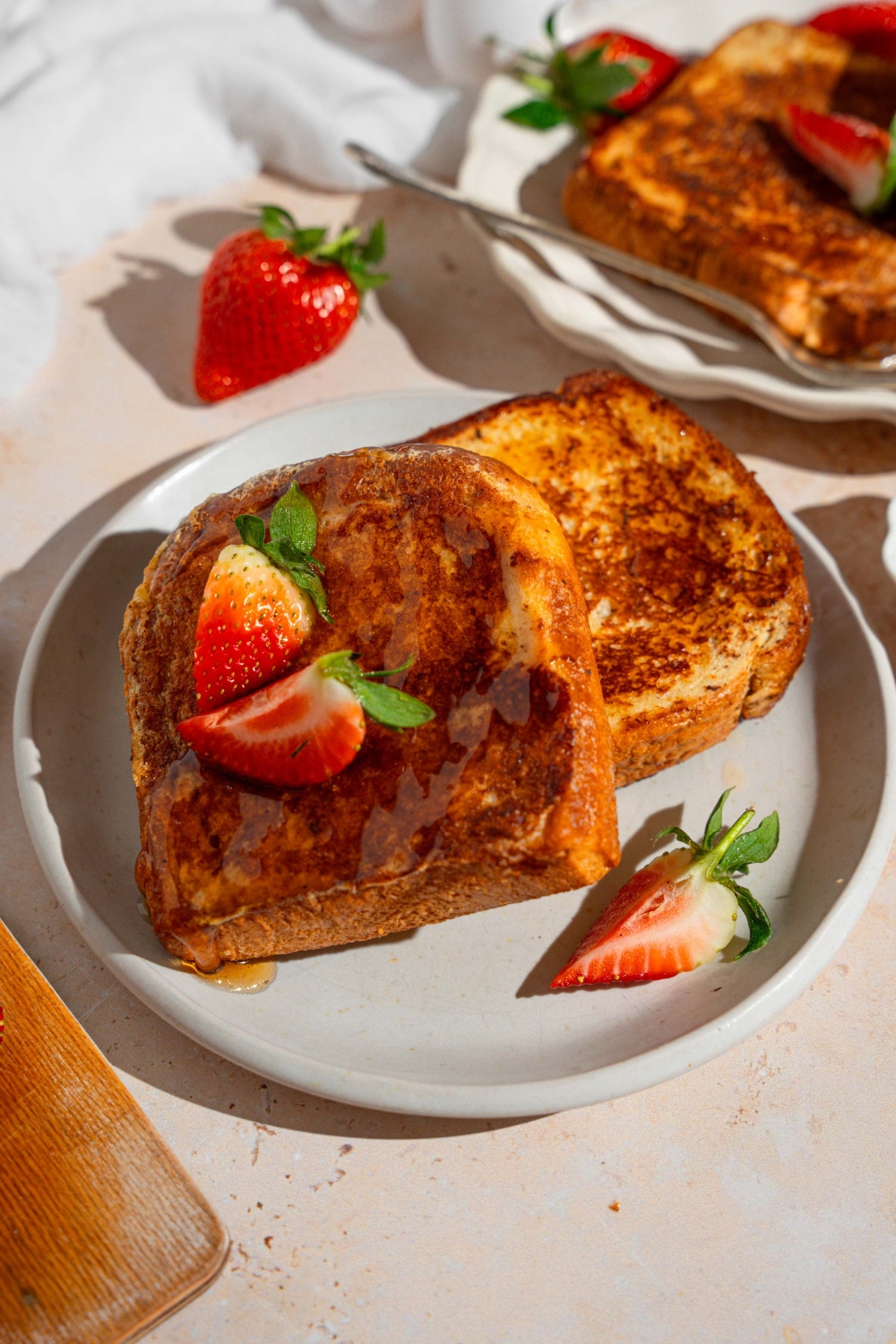 A white plate with slices of French toast drizzled with maple syrup and garnished with sliced strawberries. The plate is on a tan counter with a platter of french toast.