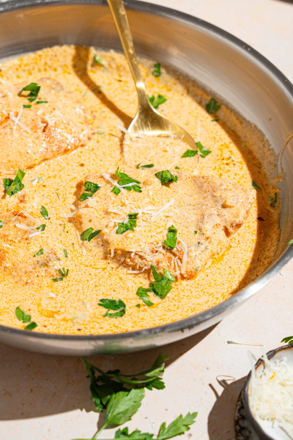 A skillet with a spoon stirring chicken breasts cooked in cajun alfredo sauce garnished with fresh parsley. The skillet is on a tan counter.