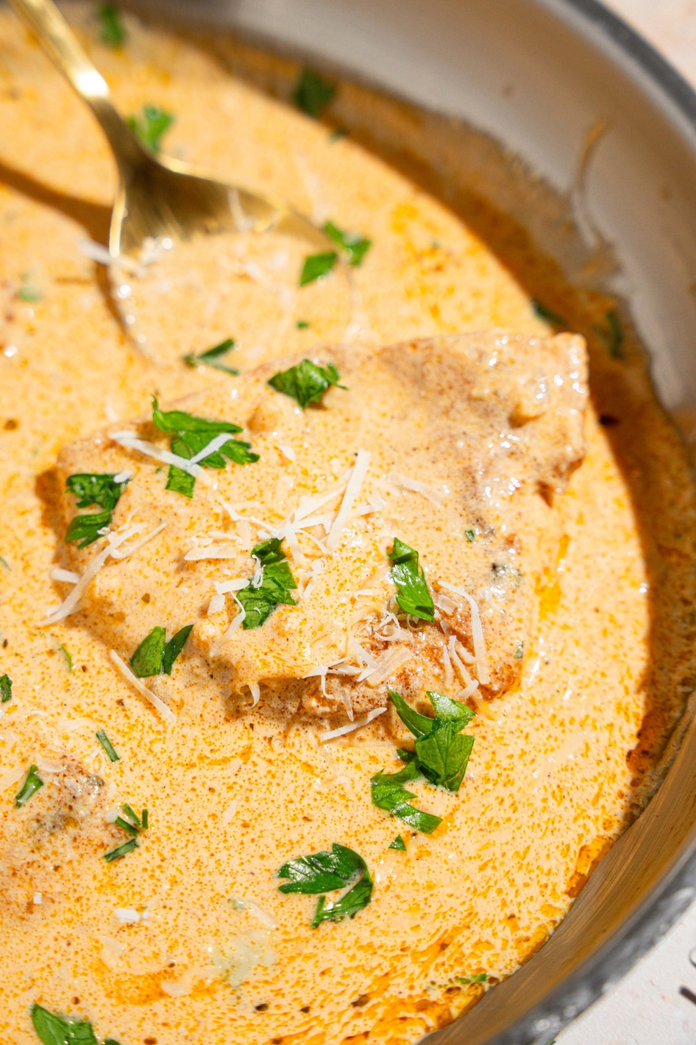A skillet with a spoon stirring chicken breasts cooked in cajun alfredo sauce garnished with fresh parsley. The skillet is on a tan counter.