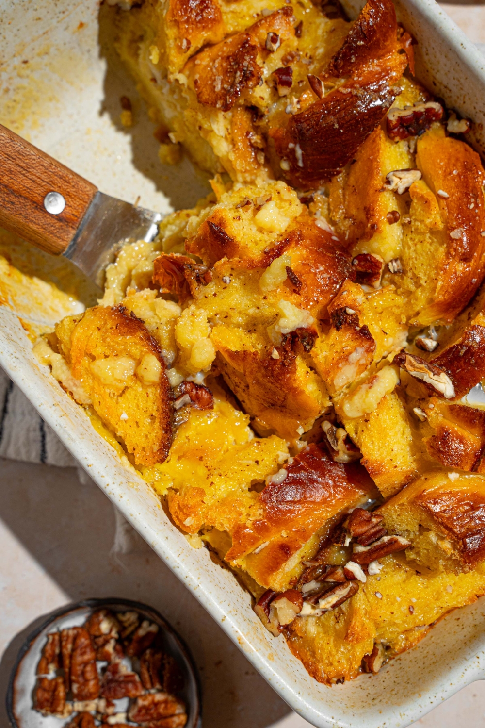 A baking dish with challah french toast with a spatula removing a piece. The dish is on a tan counter with a white striped napkin and small plate of pecans.