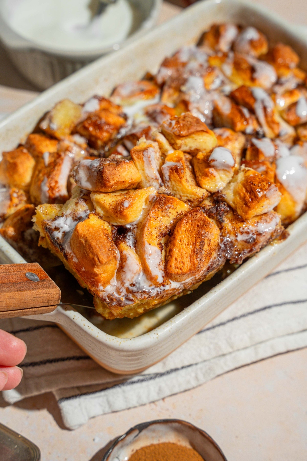 A baking dish with cinnamon roll french toast drizzled with icing. The dish is on a tan counter on a white striped napkin. A serving spatula is removing a slice from the dish.