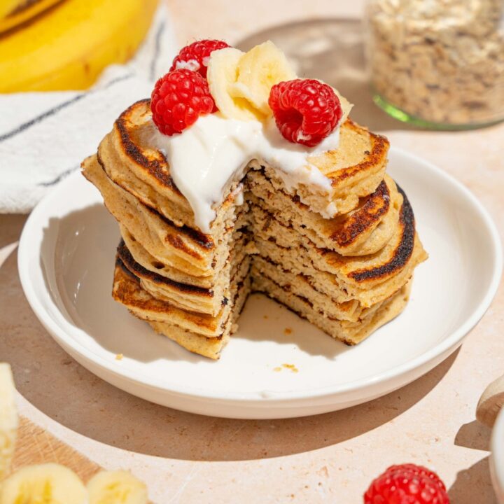 A white plate with a stack of cottage cheese banana pancakes topped with whipped cream, sliced banana, and raspberries. There is a bite taken from the stack. The plate is on a tan counter with bananas and a bowl of raspberries.