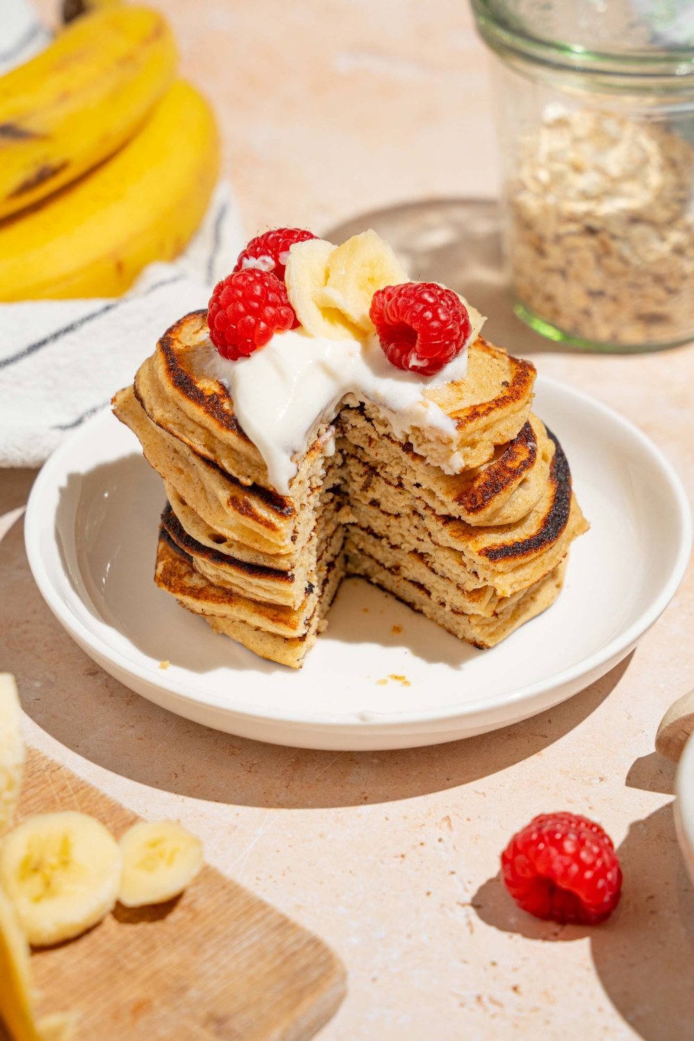 A white plate with a stack of cottage cheese banana pancakes topped with whipped cream, sliced banana, and raspberries. There is a bite taken from the stack. The plate is on a tan counter with bananas and a bowl of raspberries.